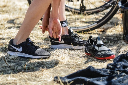 Twee benen waarbij iemand een hardloopschoen aantrekt. Op de achtergrond een fiets en op de voorgrond een fietsschoen.